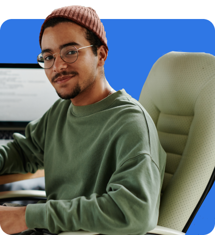 A young man wearing glasses and a beanie sits on an office chair, looking at the camera. A computer monitor with visible code hints at his tech-savvy nature. The digitally added blue background complements the scene, subtly evoking thoughts of cyber insurance and digital security.