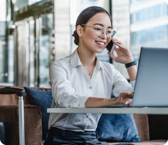 A woman wearing glasses and a white shirt sits in front of a laptop, discussing scaffolding insurance on her smartphone, and smiling. She is in a modern office environment.