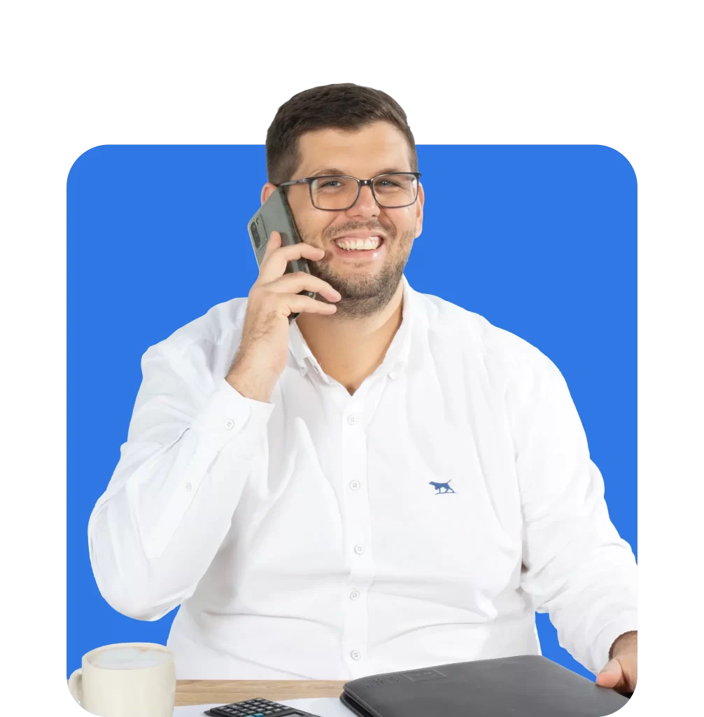 A man wearing glasses and a white shirt sits at a desk, smiling while talking on the phone. As a business insurance broker, he keeps a coffee cup, calculator, and folder close at hand to ensure all client information is accurately managed.