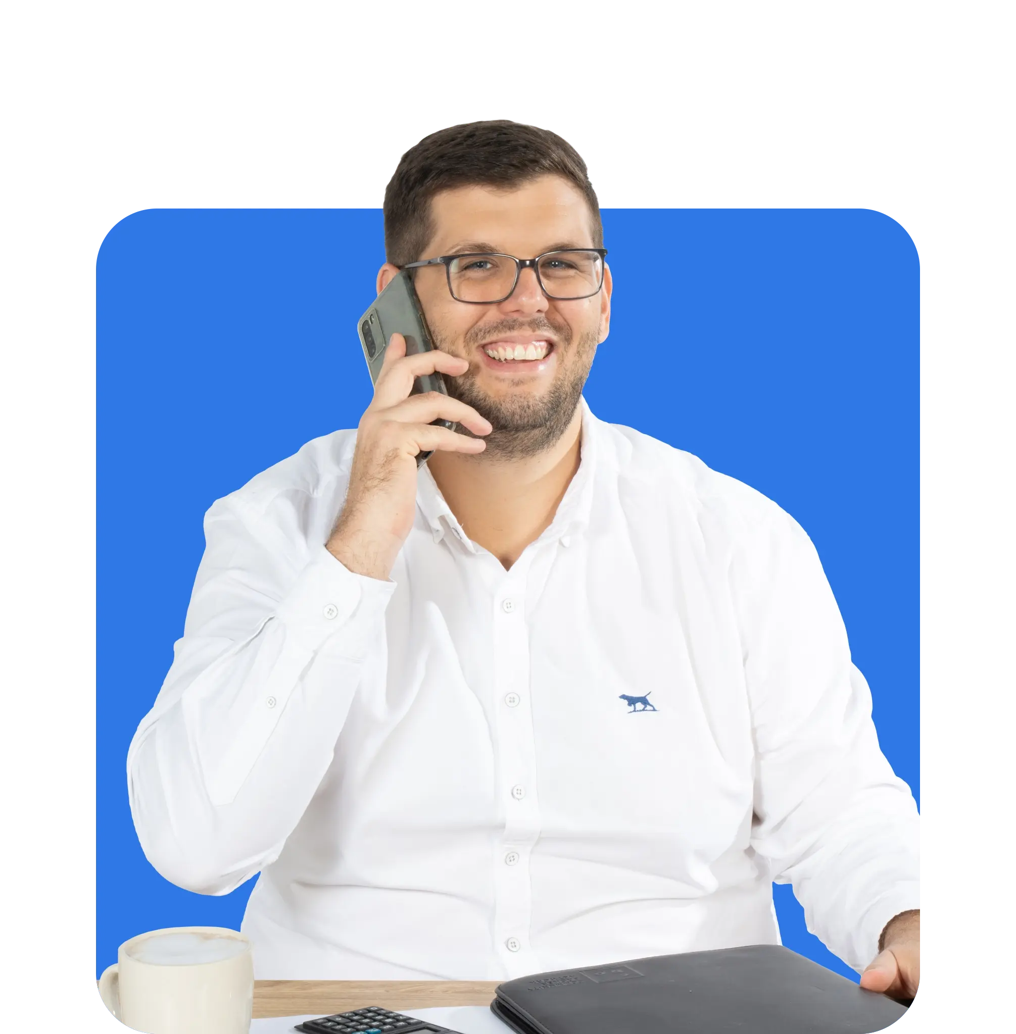 A man wearing glasses and a white shirt sits at a desk, smiling while talking on the phone. As a business insurance broker, he keeps a coffee cup, calculator, and folder close at hand to ensure all client information is accurately managed.