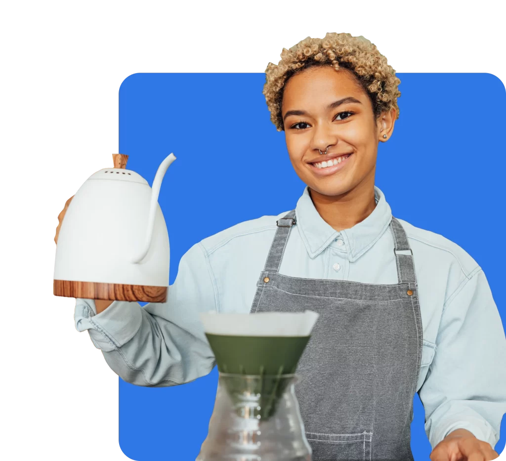 A person in a gray apron and light blue shirt is smiling while holding a white kettle, preparing a pour-over coffee against a blue background, all under the peace of mind that Public Liability Insurance brings.