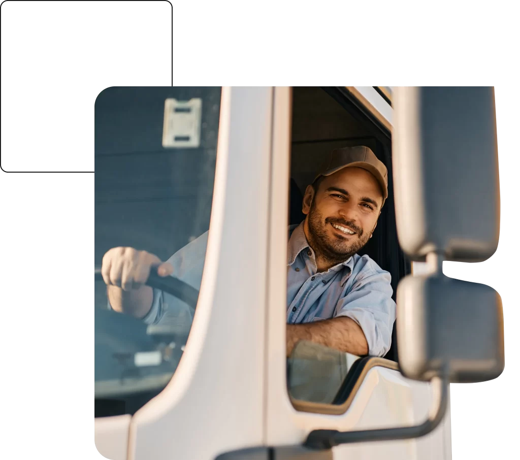 A man wearing a cap is sitting at the steering wheel inside a truck and smiling.