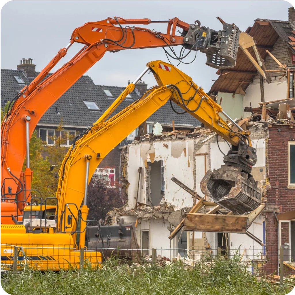 Two excavators, one orange and one yellow, demolish a multi-story brick building. Debris and partially demolished walls are visible, underscoring the importance of Public Liability Insurance on such projects.