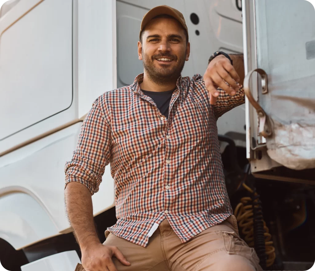 Man in a plaid shirt and cap smiles while leaning on a truck, with one foot resting on its step.