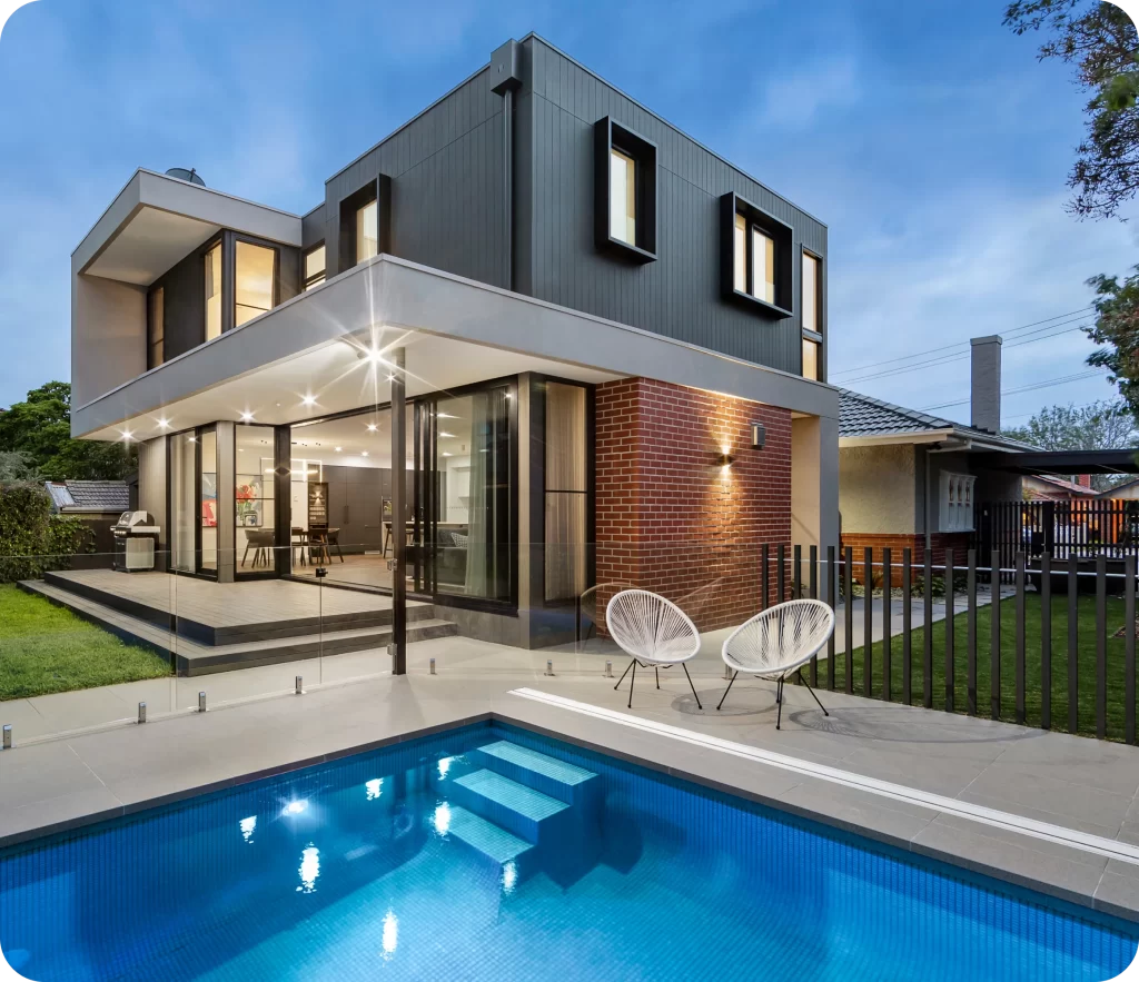 Modern two-story house with large windows, brick and metal siding, evening lighting, outdoor pool, two white chairs on a patio, and a green lawn fenced by glass and metal railings. With Prestige Home Insurance, your elegant property is always protected.