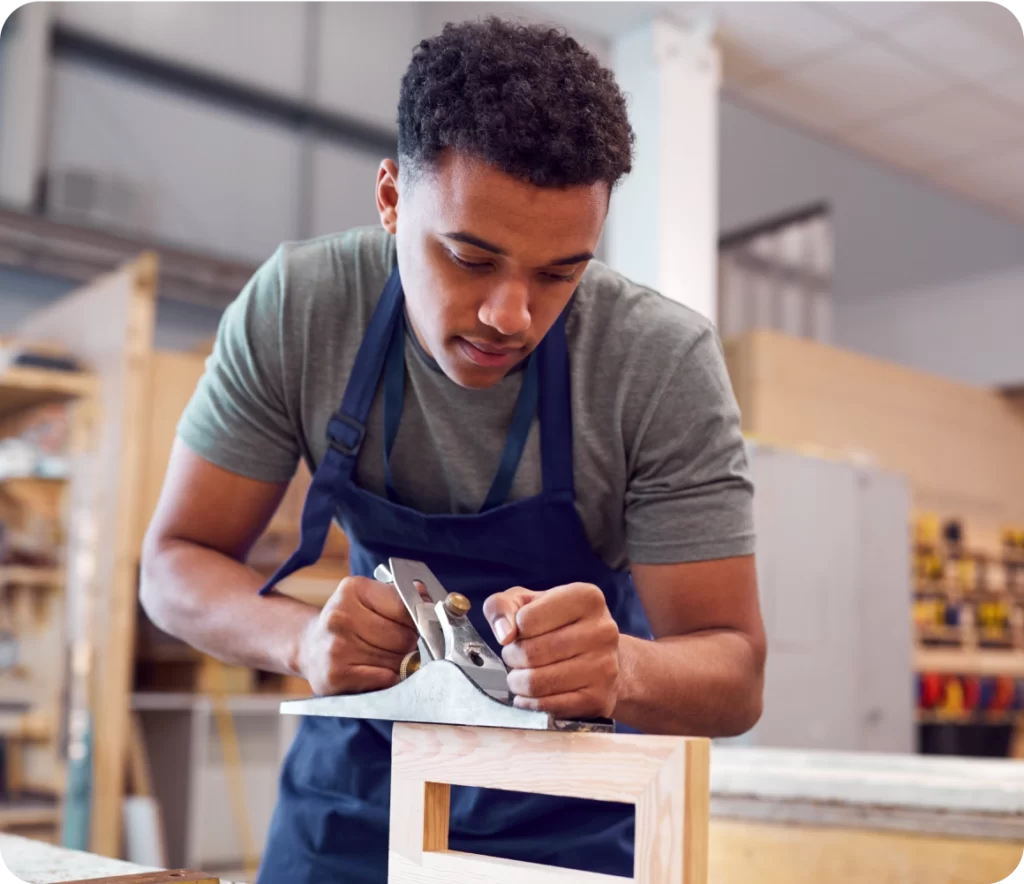 A man wearing a gray shirt and navy apron is using a hand planer to smooth a wooden piece in a workshop environment.