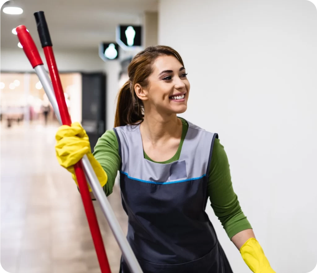 A woman in a cleaning uniform and yellow gloves holds a mop and broom, smiling. She is in a hallway with restroom signs visible in the background.