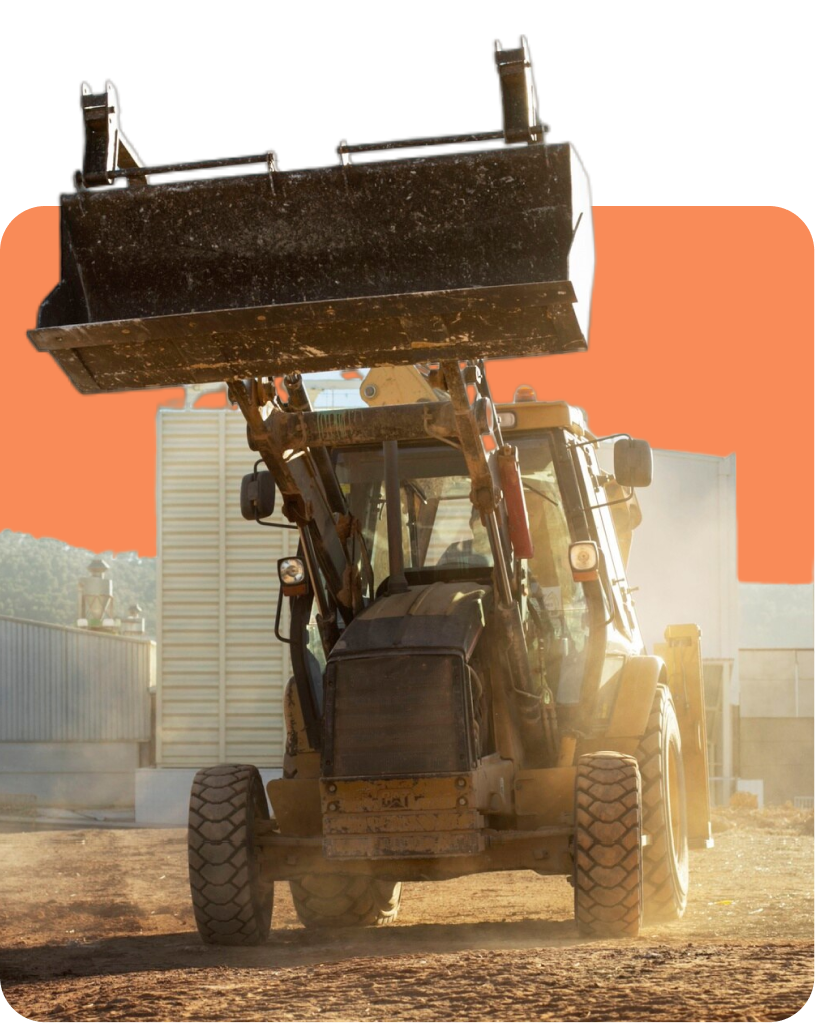 Front view of a bulldozer with its blade raised, lifting dirt at a construction site with industrial buildings in the background.