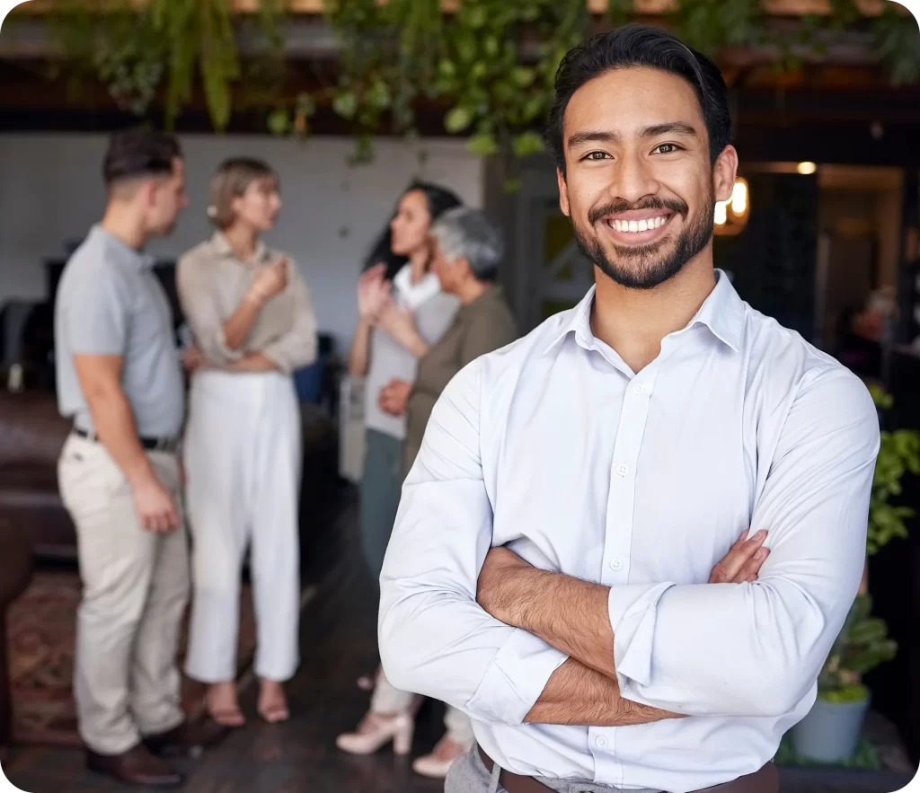 A man with crossed arms stands in the foreground smiling, while a group of four people discuss Public Liability Insurance in the background in a room adorned with plants.