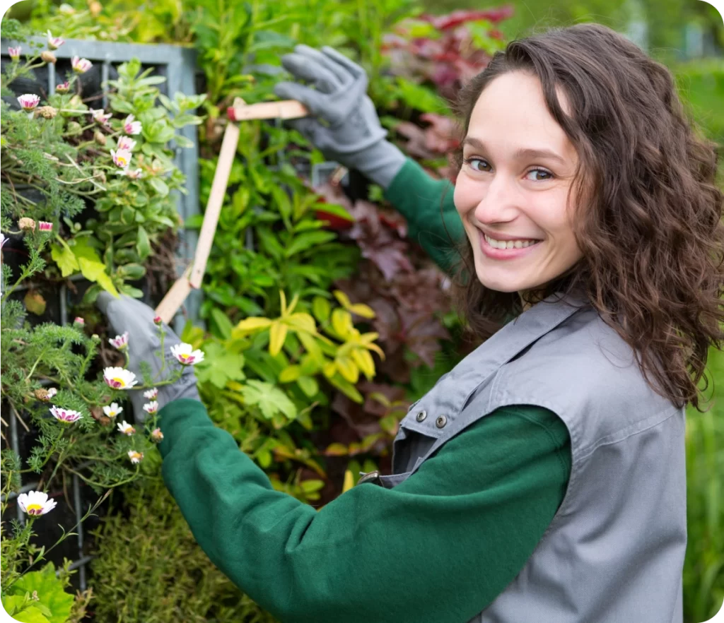 A woman wearing gardening gloves and a vest smiles at the camera while trimming plants on a vertical garden.