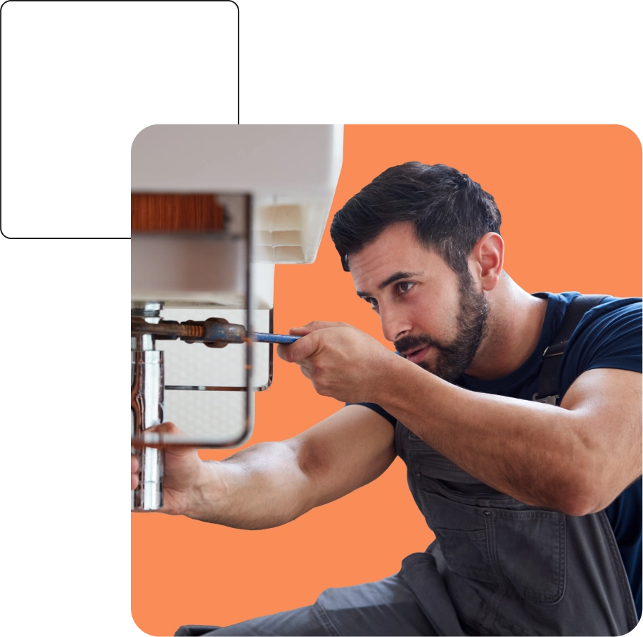 A man wearing overalls is using a screwdriver to repair a pipe under a sink against an orange background.