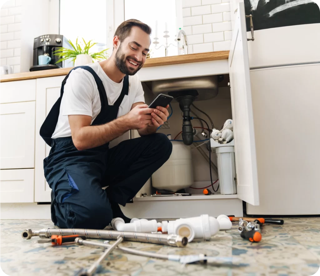 A smiling plumber in blue overalls and a white shirt kneels near a kitchen sink, looking at his phone. Plumbing tools and pipes are spread out on the floor in front of him.