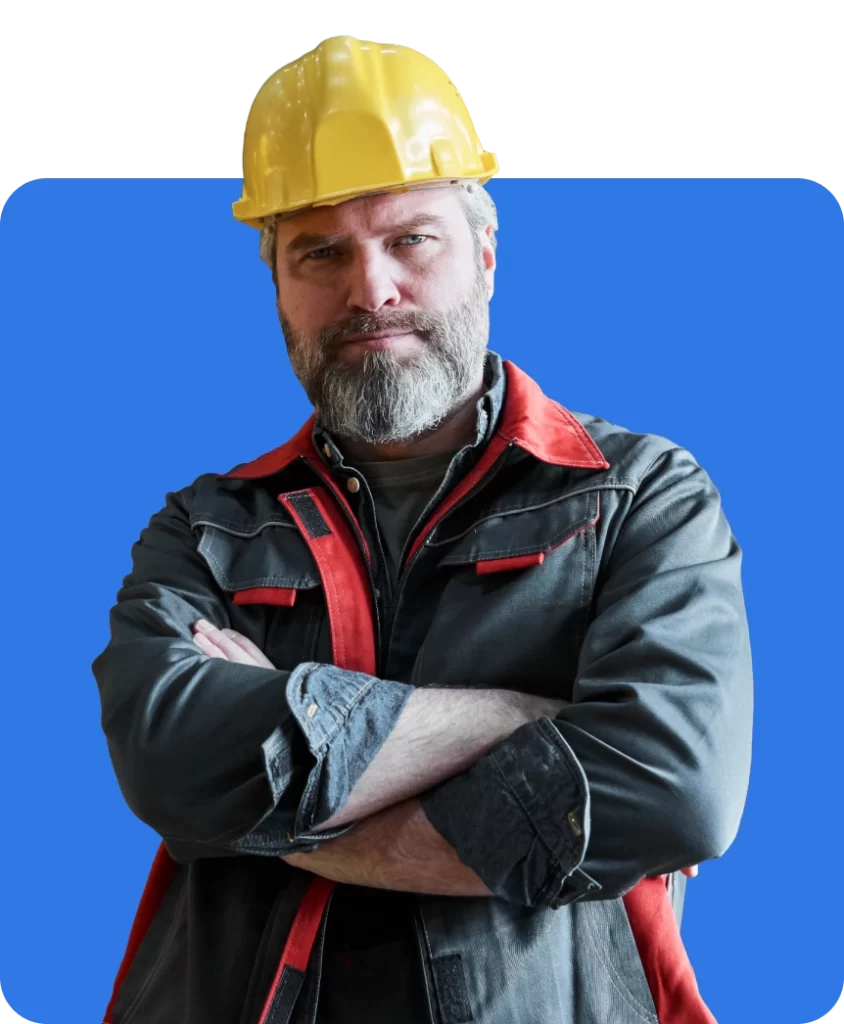 A man with a beard wearing a yellow hard hat and black jacket with red accents stands against a blue background, arms crossed.