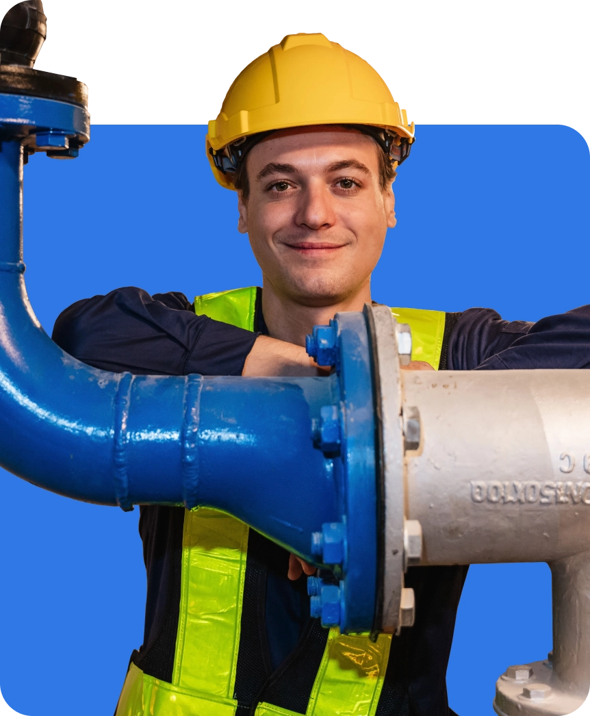 A person wearing a yellow hard hat and high-visibility vest stands next to large blue and white industrial pipes.